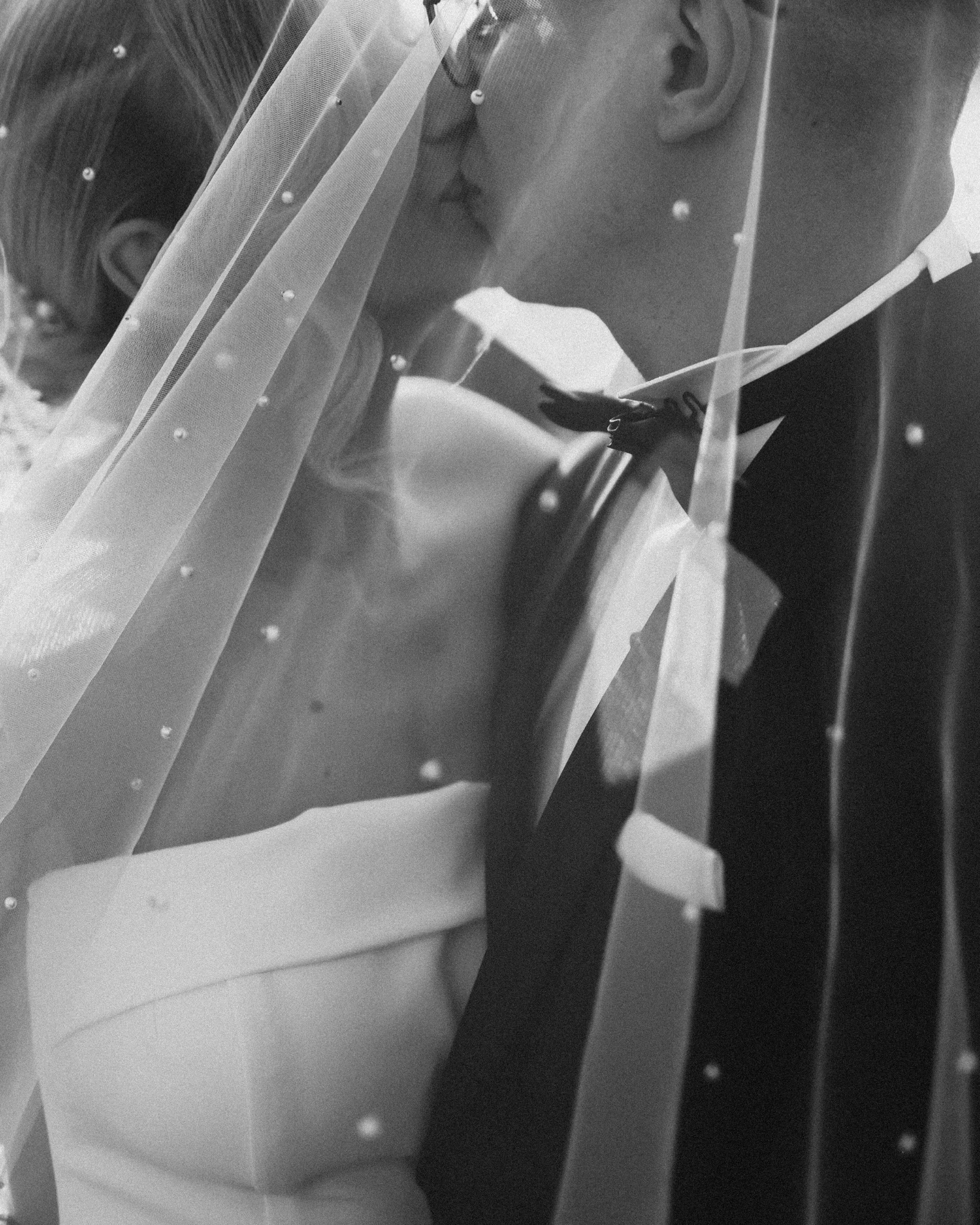 Czarno-białe ujęcie całujących się nowożeńców, przesłoniętych welonem z delikatnymi zdobieniami, z widocznym fragmentem sukni i garnituru.