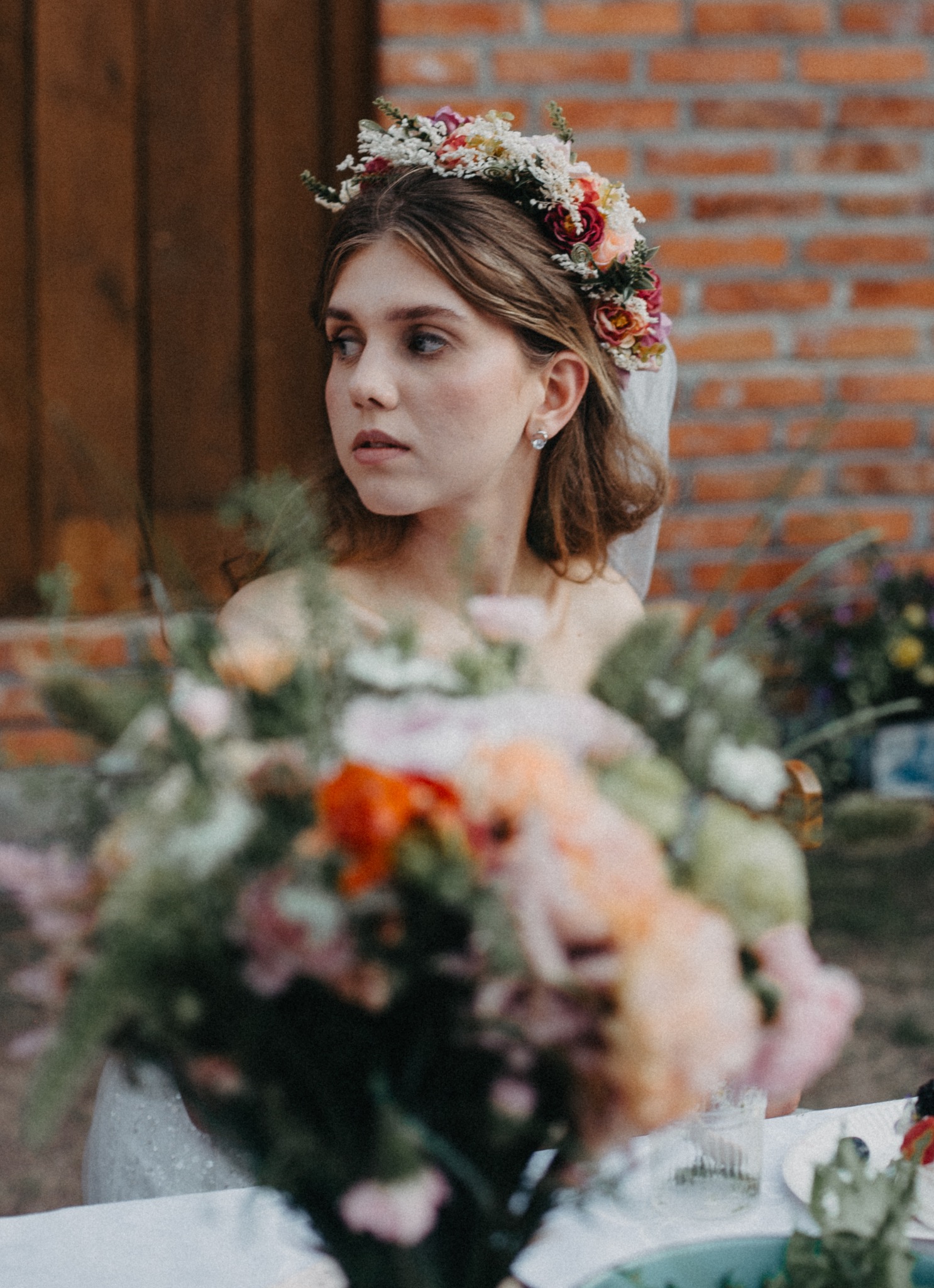 Panna młoda z wiankiem we włosach spogląda w bok, Wilkasy. W tle ceglana ściana i bukiet kwiatów na stole. Subtelny makijaż i delikatna biżuteria.