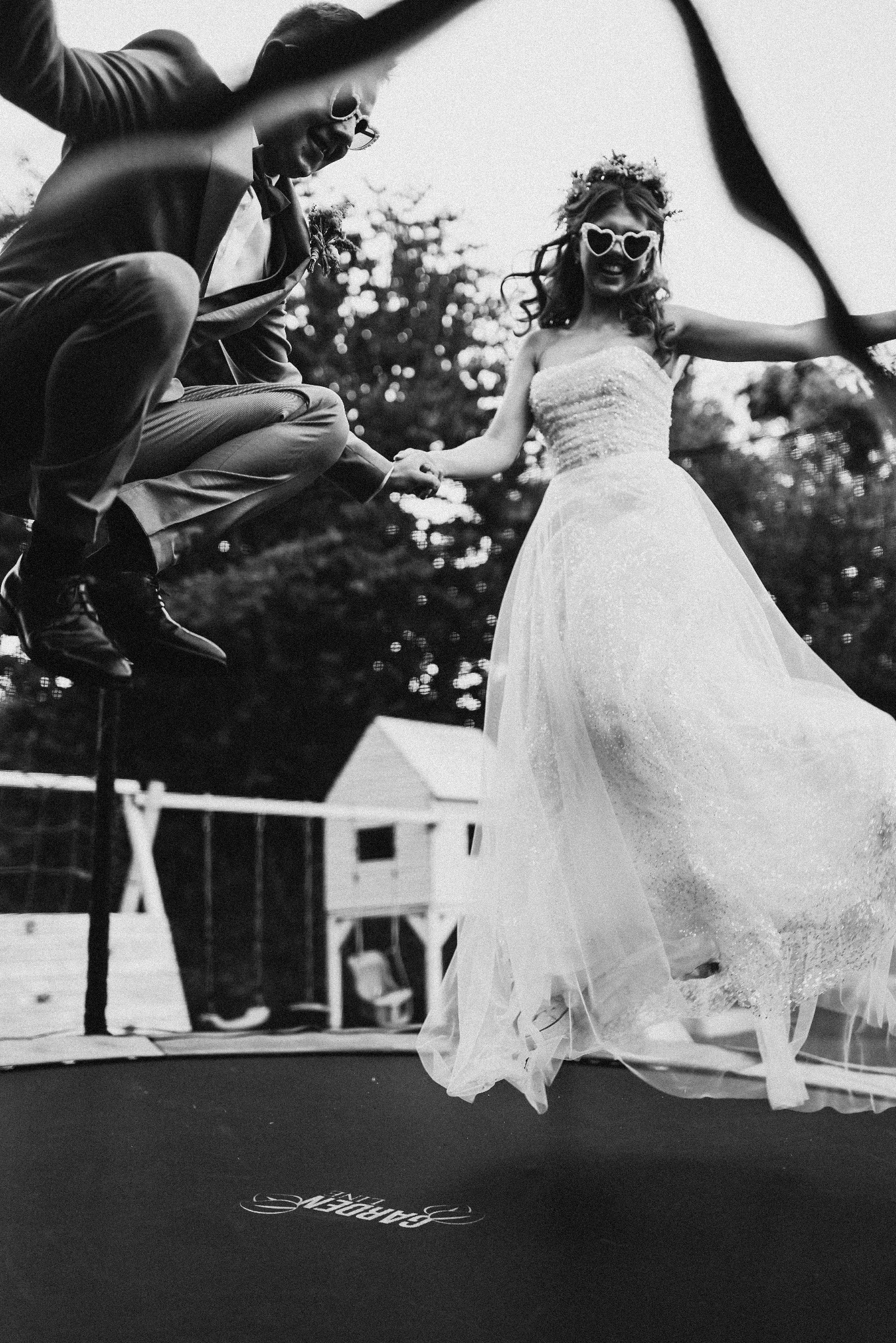 Czarno-białe zdjęcie pary młodej skaczącej na trampolinie w okularach w kształcie serc. Ona w sukni ślubnej, on w garniturze. W tle drzewa i mały domek.