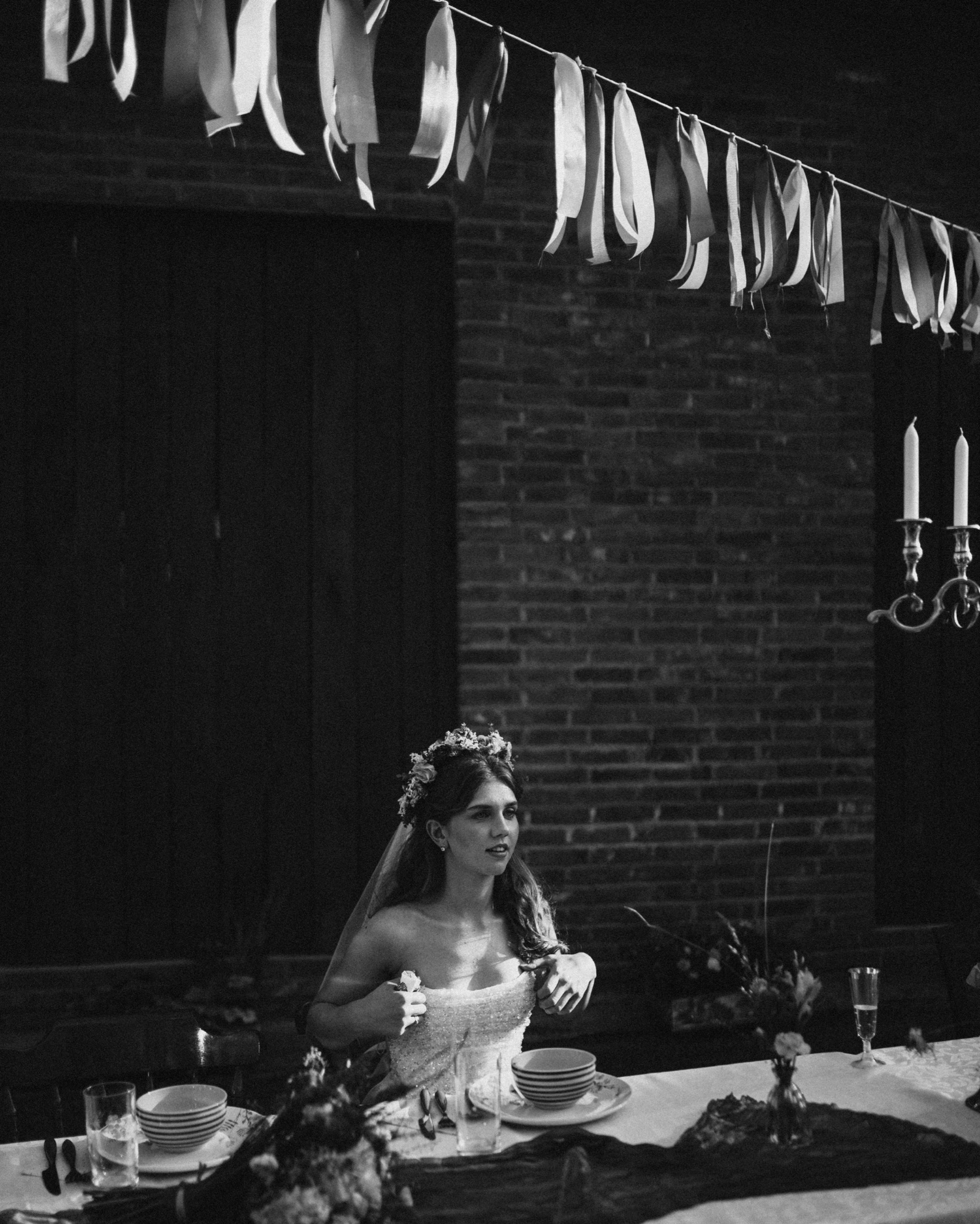 Czarno-białe zdjęcie panny młodej siedzącej przy stole udekorowanym kwiatami i świecami, w tle ceglana ściana i girlanda z wstążek.