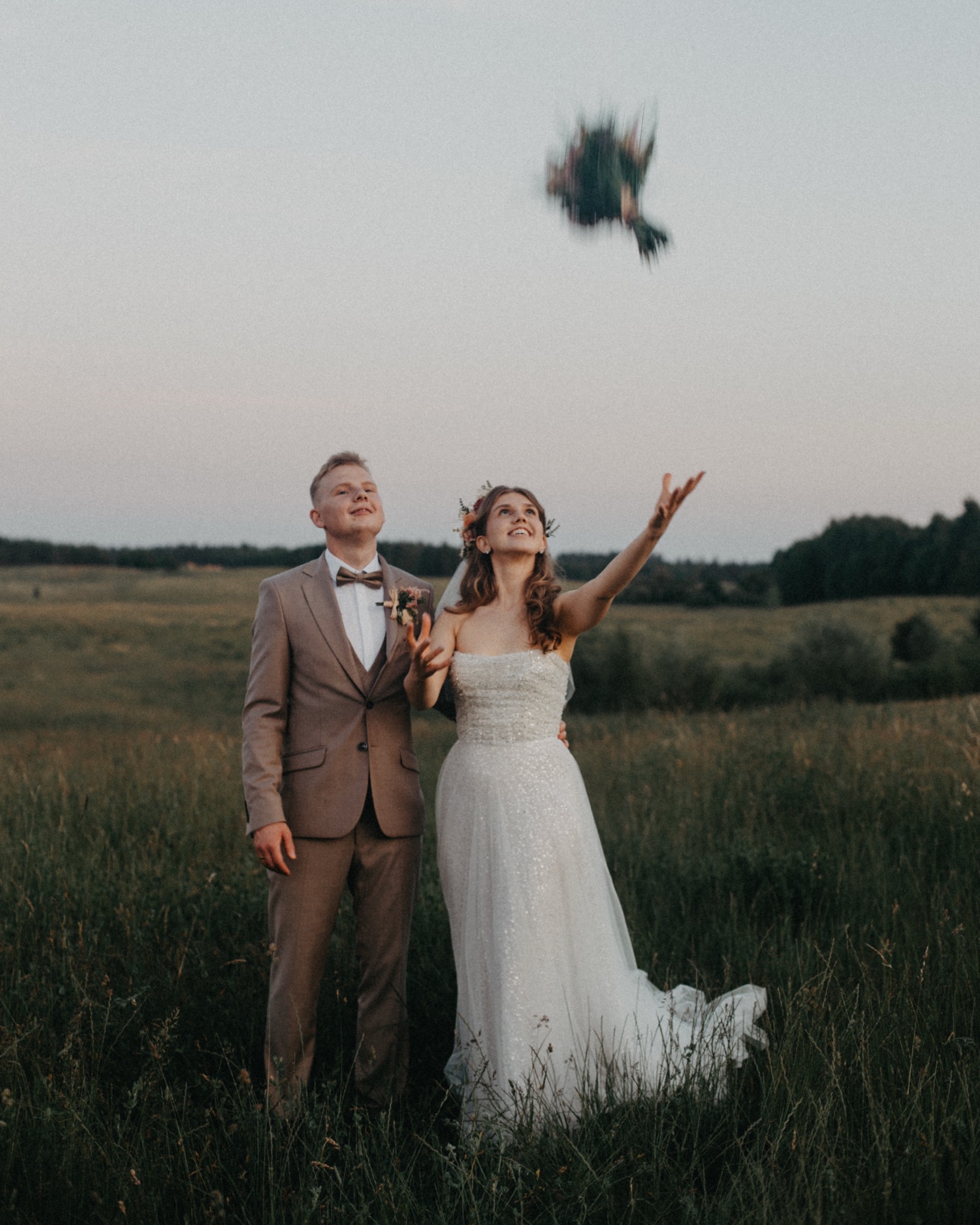 Para młoda w plenerze, panna młoda rzuca bukiet w górę. On w brązowym garniturze, ona w sukni z cekinami. Trawa i drzewa w tle, niebo szare, wieczorny klimat.