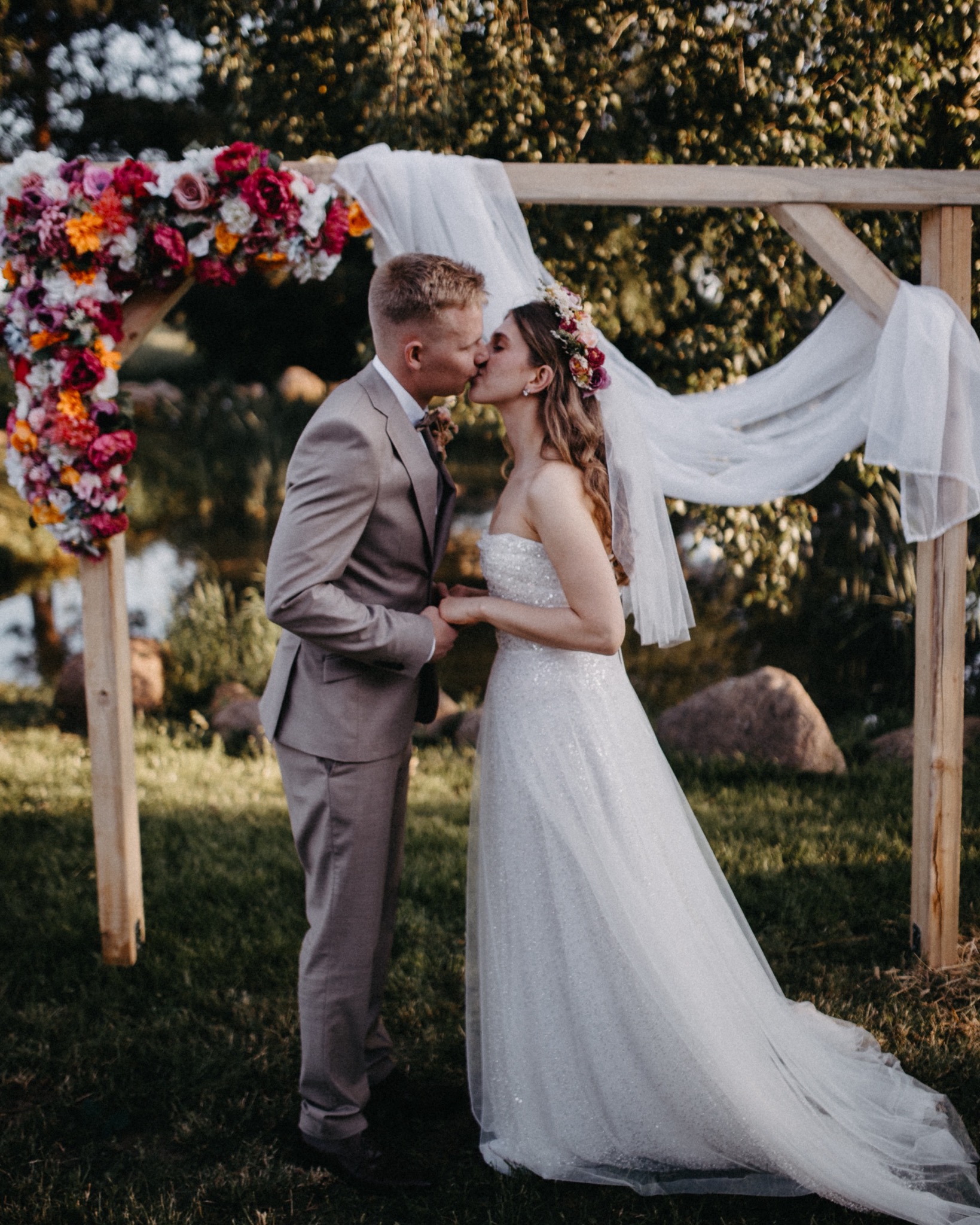 Para młoda całuje się pod drewnianym łukiem udekorowanym kwiatami i białym materiałem, na tle zieleni i wody. Ujęcie w plenerze, w ciepłych barwach.