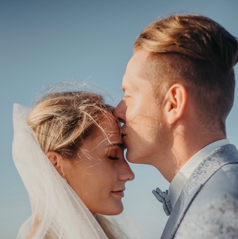 Młoda para: on w popielatym garniturze, ona w welonie. Delikatny pocałunek w czoło na tle błękitnego nieba. Romantyczna chwila uwieczniona w kadrze.