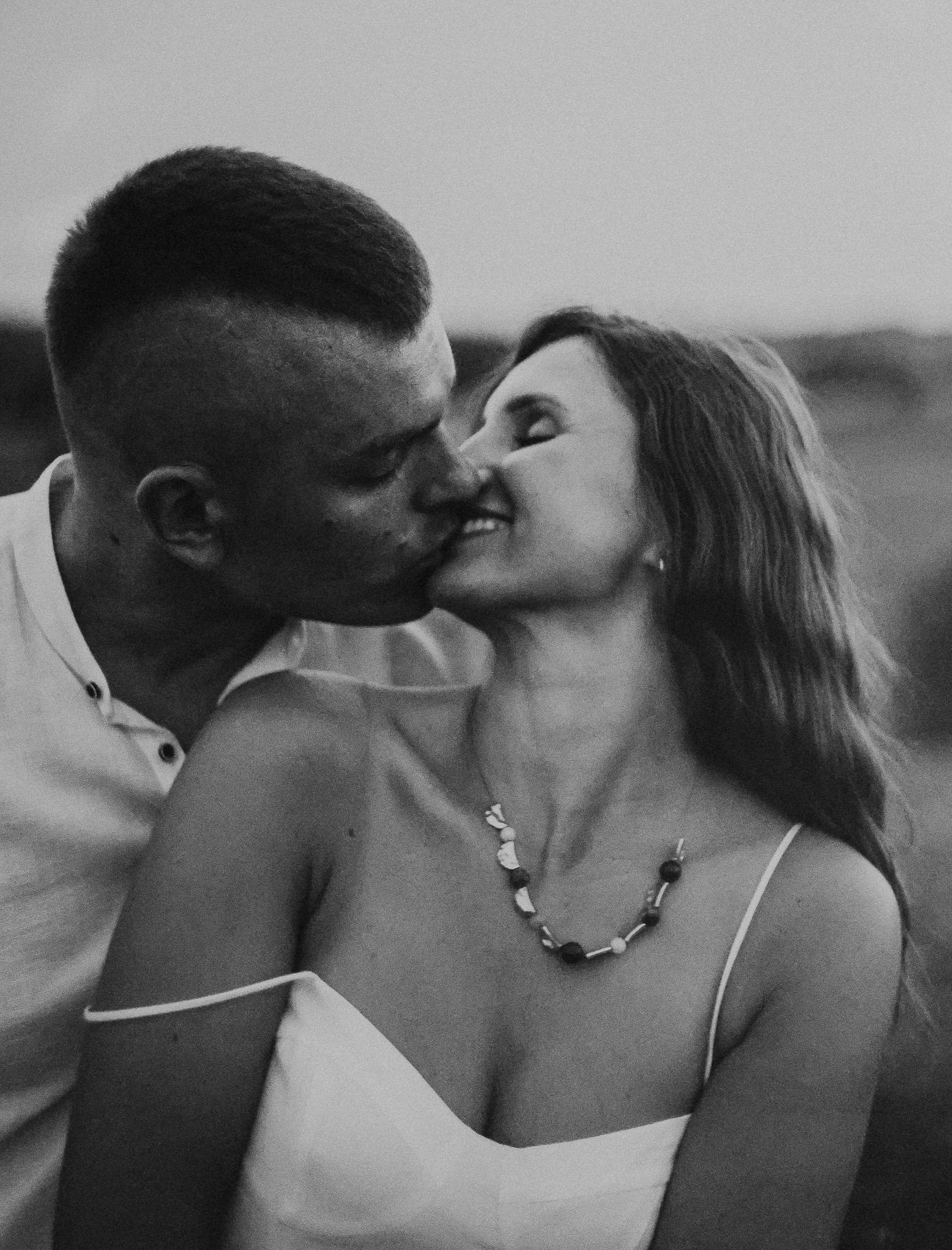 Czarno-białe zbliżenie całującej się pary. Kobieta z naszyjnikiem i sukienką na ramiączkach, mężczyzna w koszuli z rozpiętym kołnierzykiem. Romantyczny moment uchwycony w plenerze.