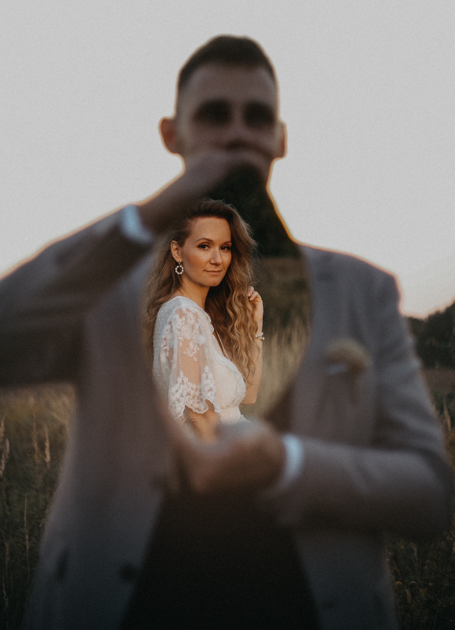 Kreatywne ujęcie pary: kobieta w koronkowej sukience na pierwszym planie, mężczyzna w beżowym garniturze w tle, z efektem głębi i naturalnym oświetleniem.