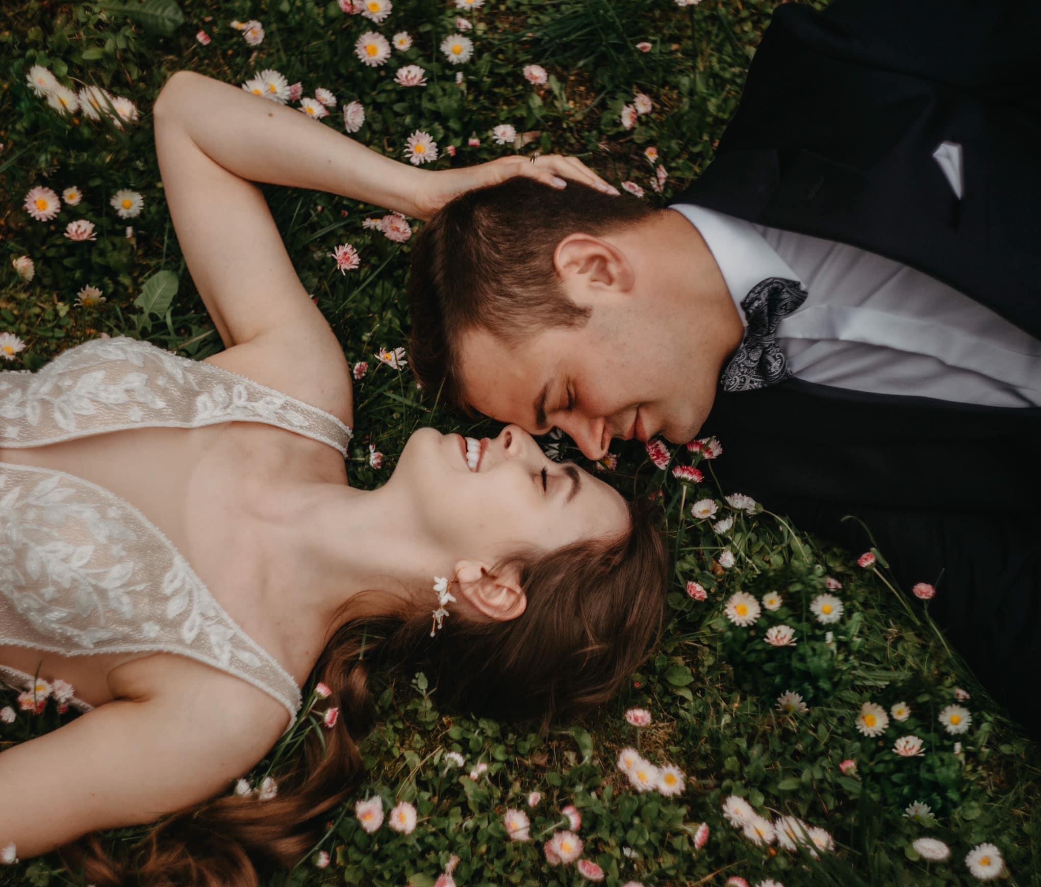 Para młoda leży na trawie wśród stokrotek. Panna młoda w sukni z koronkowymi aplikacjami, pan młody w czarnym garniturze z muszką. Romantyczna sesja w plenerze.