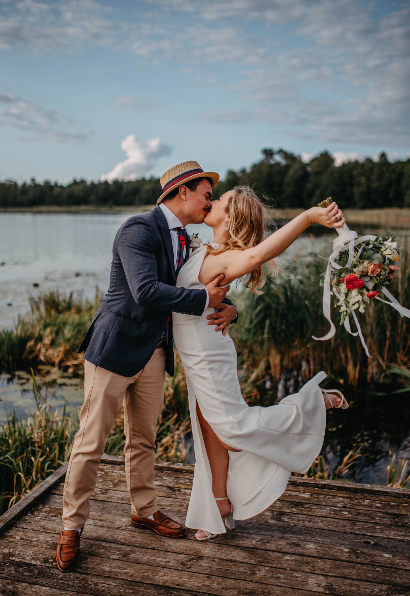 Para młoda całuje się na drewnianym pomoście nad jeziorem. Panna młoda w białej sukni z bukietem, pan młody w garniturze i kapeluszu.