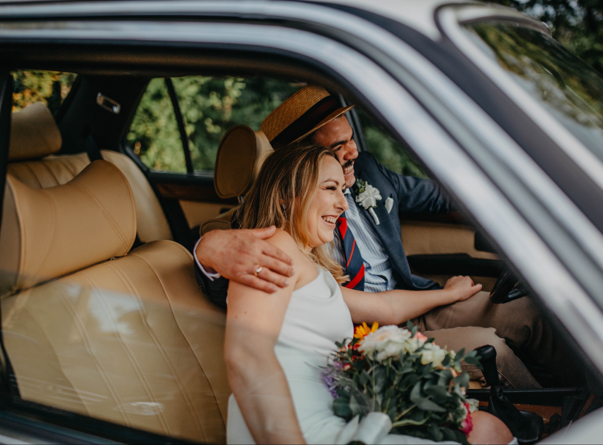 Uśmiechnięta para młoda w samochodzie z bukietem. Pan młody w kapeluszu i marynarce, panna młoda w białej sukience. Jasne wnętrze auta, widok z boku przez szybę.