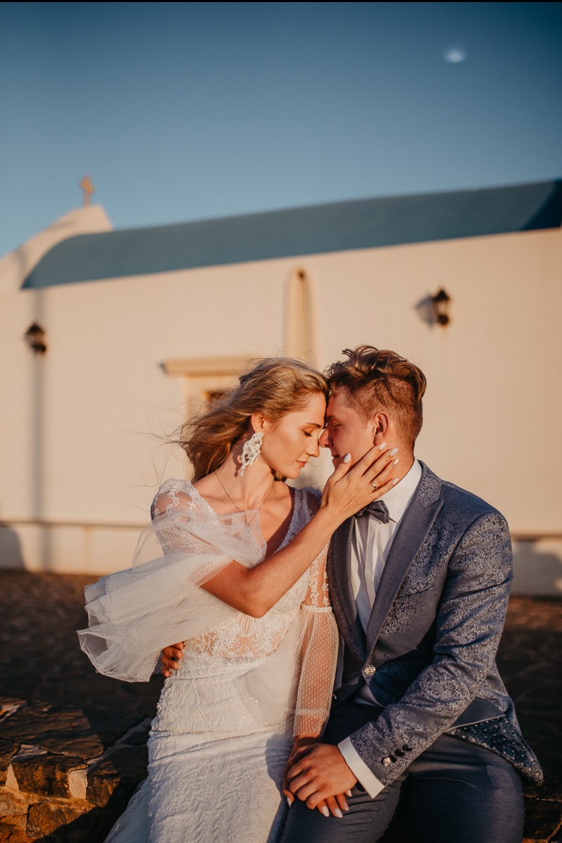 Para młoda w objęciach na tle białego kościoła z niebieskim dachem. Kobieta w koronkowej sukni, mężczyzna w eleganckim garniturze. Romantyczny kadr, ciepłe światło zachodzącego słońca.