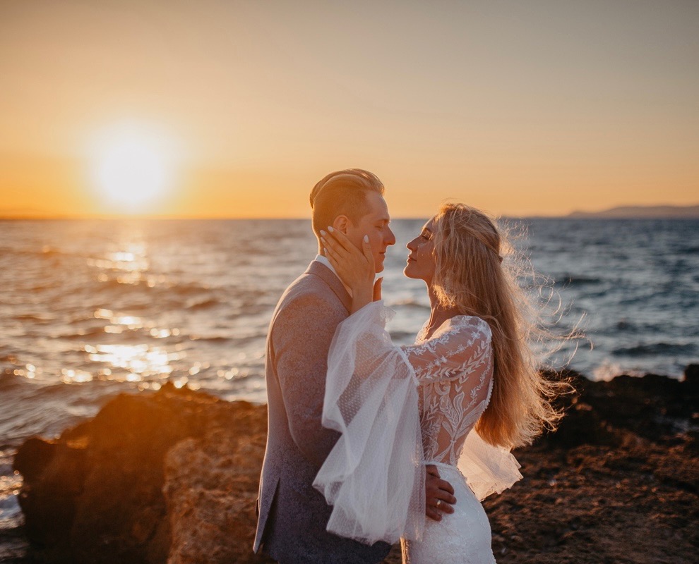 Para w objęciach na tle zachodzącego słońca nad morzem. Kobieta w koronkowej sukni, mężczyzna w garniturze. Romantyczna sceneria z ciepłym światłem.