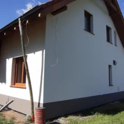 Dom jednorodzinny w trakcie prac wykończeniowych elewacji: ocieplenie części ściany z widoczną konstrukcją drewnianą, pomalowana na biało górna część elewacji i szary cokół, widoczne rury spustowe.