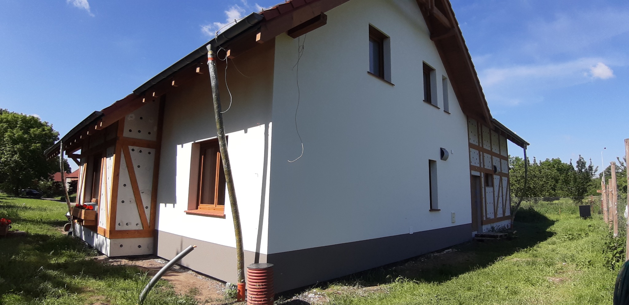 Dom jednorodzinny w trakcie prac wykończeniowych elewacji: ocieplenie części ściany z widoczną konstrukcją drewnianą, pomalowana na biało górna część elewacji i szary cokół, widoczne rury spustowe.