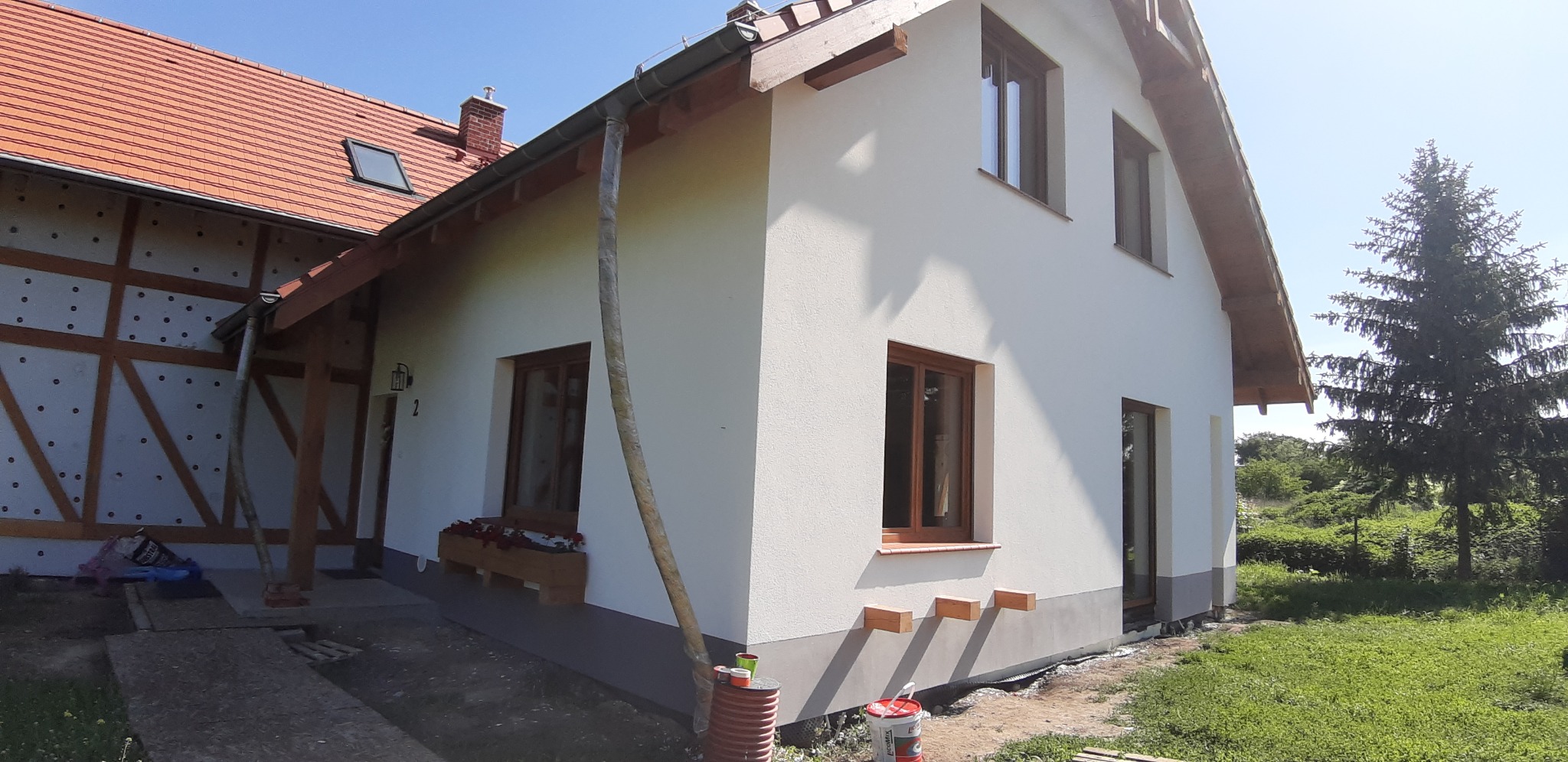 Boczny widok domu po ociepleniu i świeżym pomalowaniu elewacji na biało, z widocznymi brązowymi oknami i drewnianymi elementami wykończeniowymi, z fragmentem rusztowania w dolnej części.