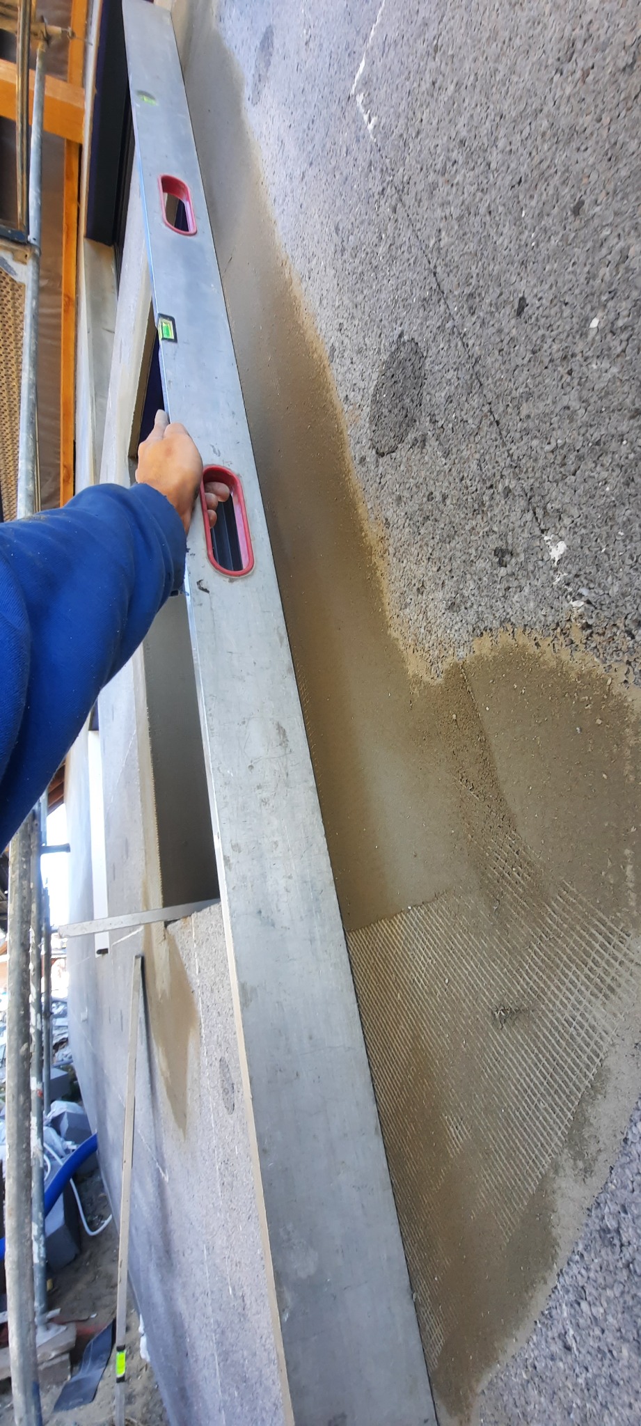 Sprawdzanie pionu ściany z ociepleniem styropianowym przy użyciu poziomicy, widoczna siatka zatopiona w kleju na elewacji.