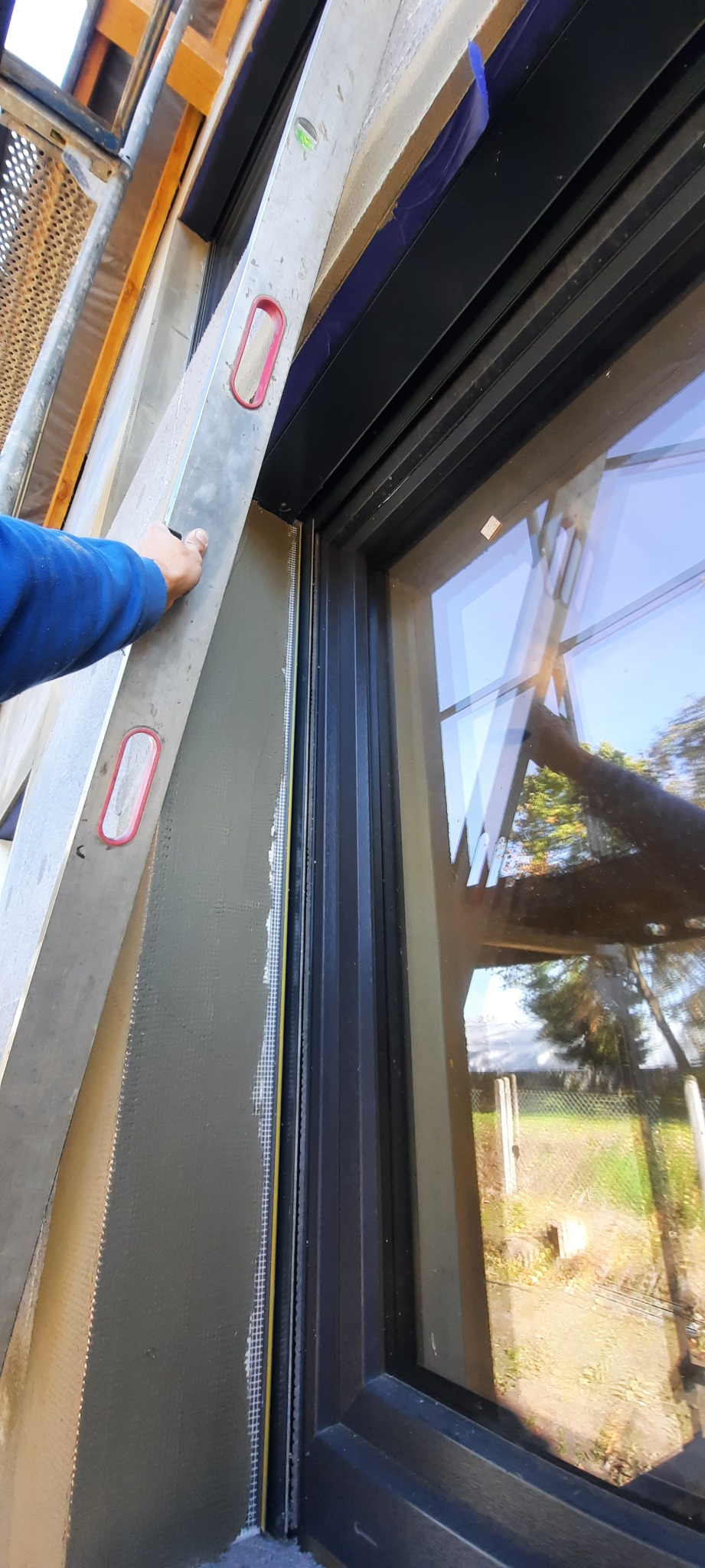 Przymocowywanie poziomicy do ocieplonej ściany budynku obok okna z czarną ramą, widoczna siatka wzmacniająca pod ociepleniem.