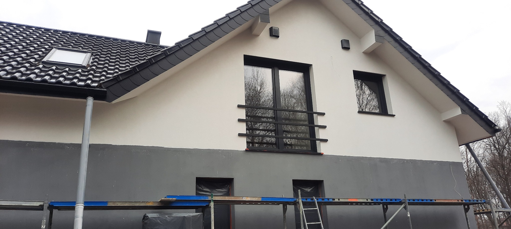 Dwukolorowa elewacja domu w Pieszycach, z szarą dolną częścią i białą górną, widoczna na rusztowaniu w trakcie prac wykończeniowych.