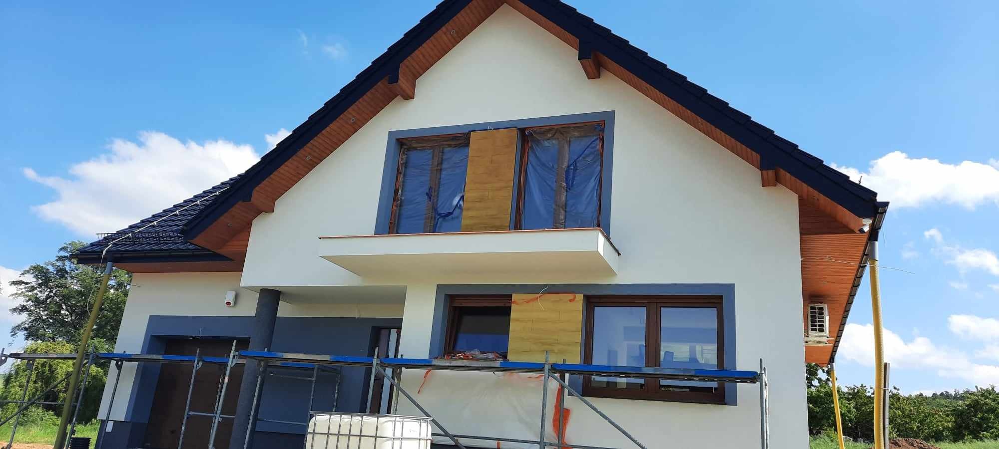 Dom jednorodzinny w trakcie prac elewacyjnych: zabezpieczone okna folią, częściowo nałożona warstwa izolacyjna, widoczne rusztowania.