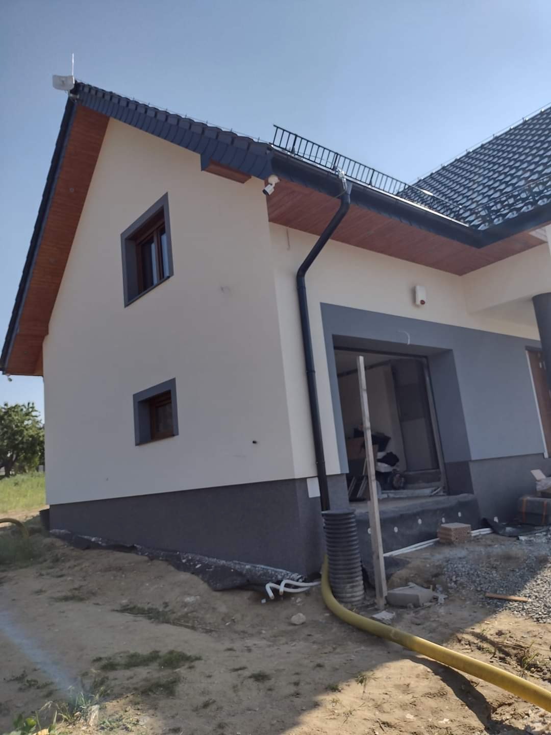 Nowo otynkowany dom jednorodzinny z ciemnoszarą podmurówką i białą elewacją górnej części, widoczne okna z ciemnymi ramami, czarna rynna i fragment dachu z ciemną dachówką, teren wokół domu...