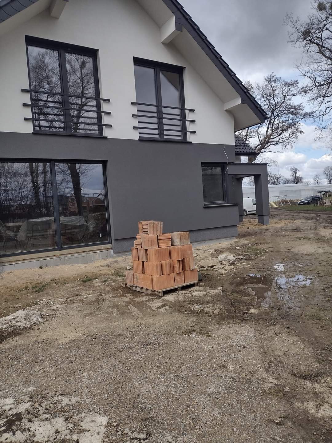 Dwukondygnacyjny dom z szarą elewacją i czarnymi oknami; sterta czerwonych cegieł na palecie przed budynkiem.