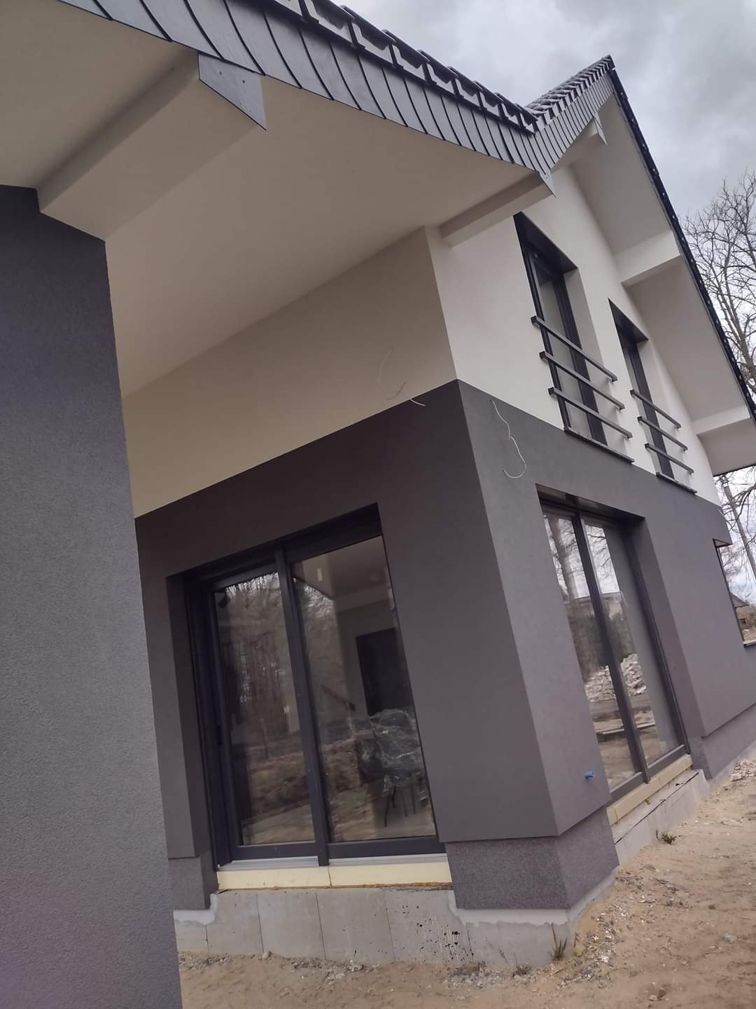 Nowoczesny dom z dwukolorową elewacją: szary dół i biała góra, widoczne okna i fragment dachu ze skośnymi dachówkami, teren wokół domu w trakcie prac wykończeniowych.