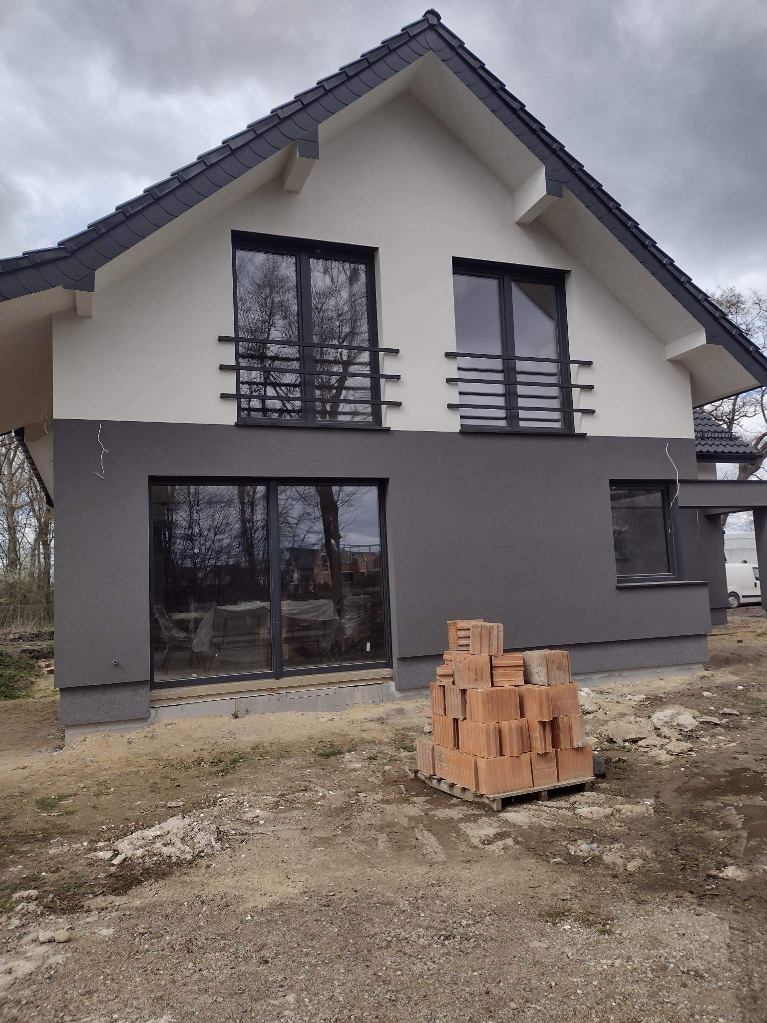 Dwukondygnacyjny dom z nową elewacją w odcieniach szarości i bieli, widoczne okna z czarnymi ramami i dekoracyjnymi poręczami, paleta z cegłami na pierwszym planie, teren wokół domu w trakcie prac...