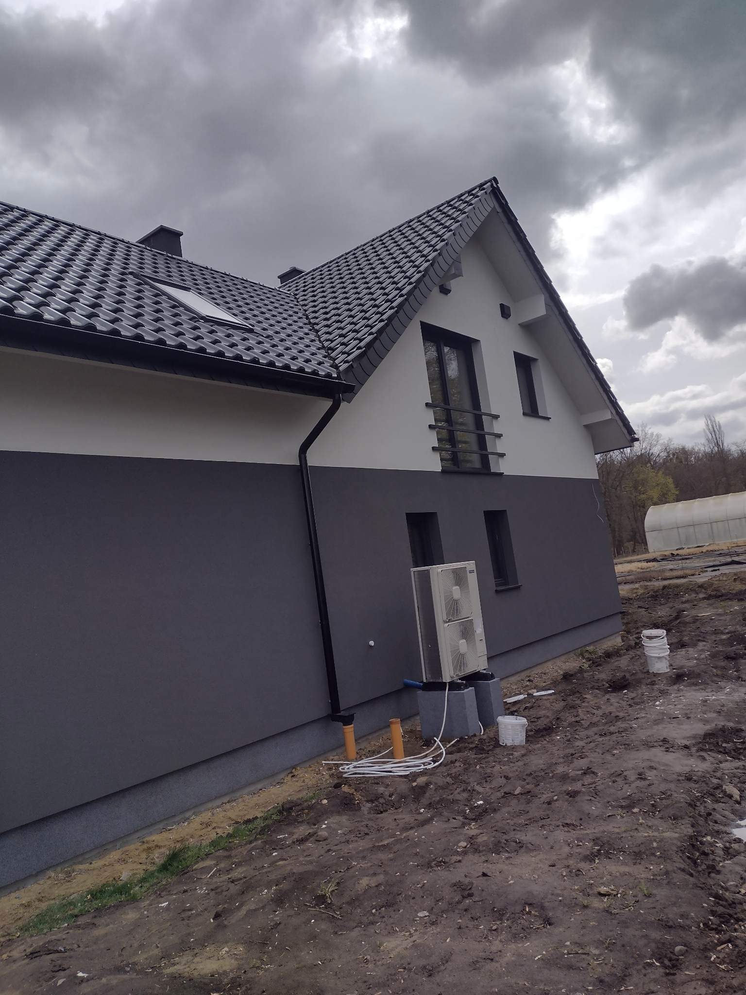 Dwukondygnacyjny dom z szarą elewacją, białą górą i ciemną dachówką, widoczny klimatyzator zewnętrzny i teren po pracach budowlanych