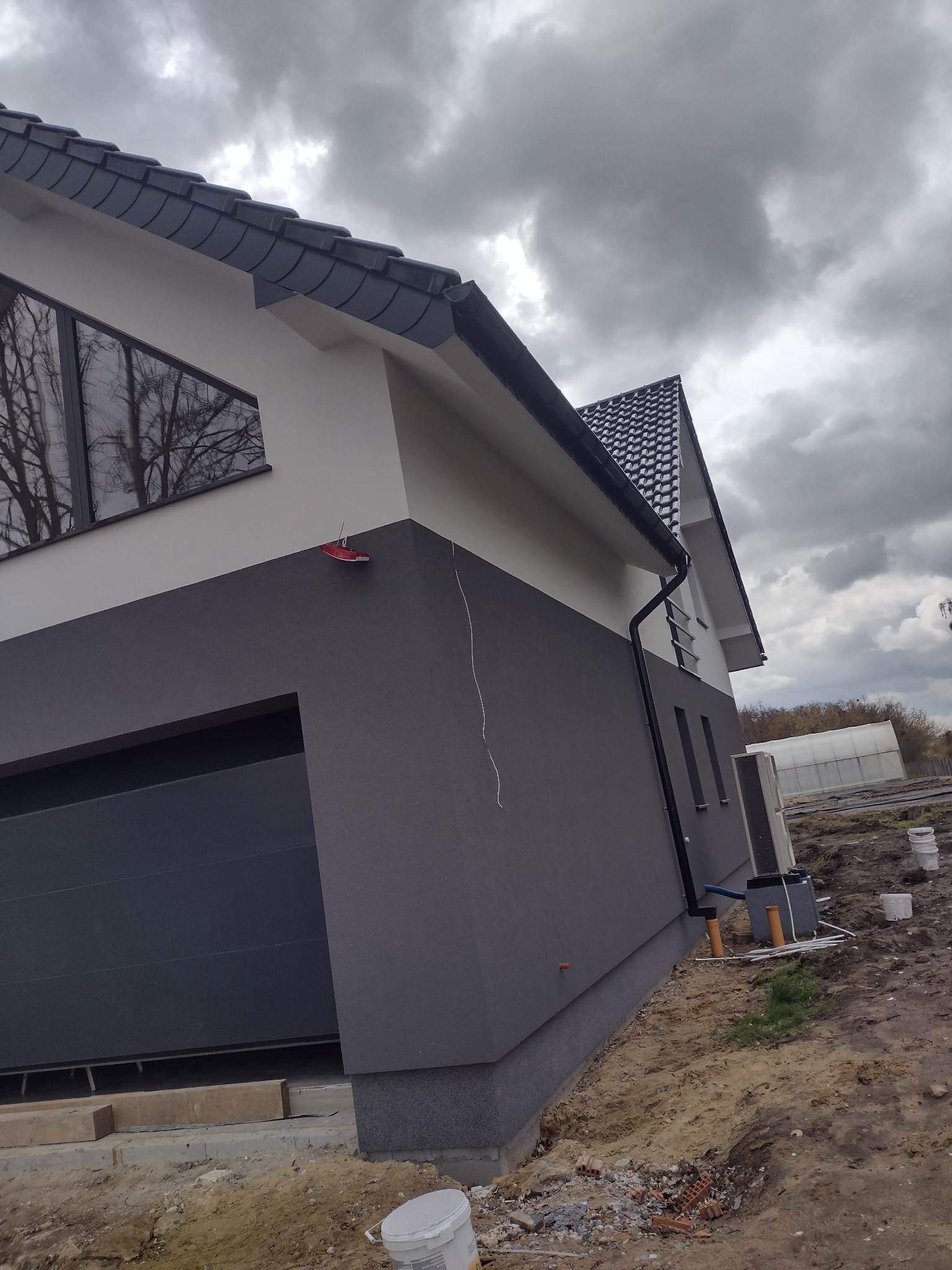 Dwukolorowa elewacja domu jednorodzinnego z szarą bramą garażową i widocznymi elementami wykończeniowymi wokół, w tle pochmurne niebo i szklarnia.