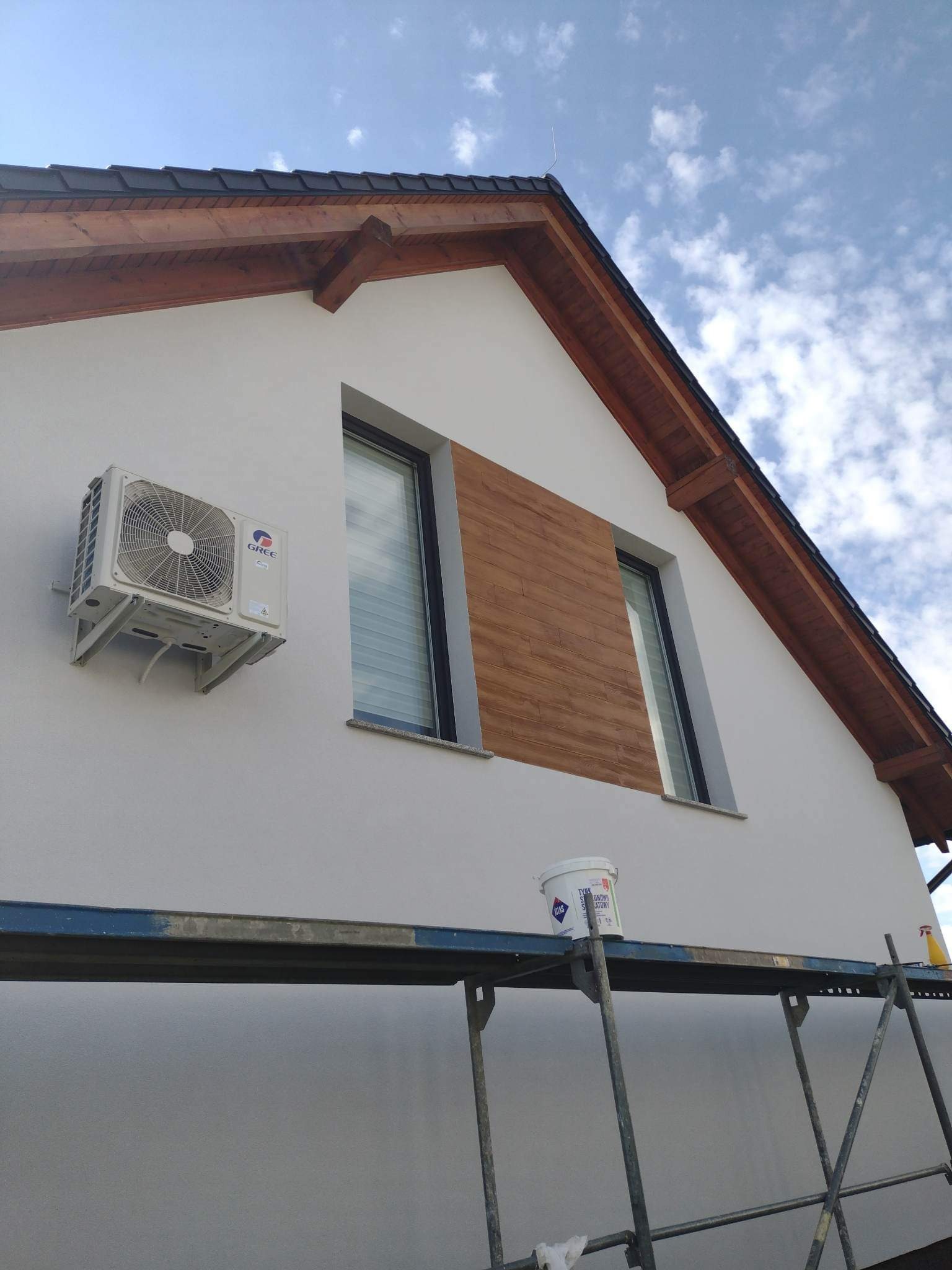 Ocieplona i pomalowana elewacja domu jednorodzinnego z widocznym klimatyzatorem, drewnianą okładziną elewacyjną, fragmentem rusztowania i wiadrem z farbą.