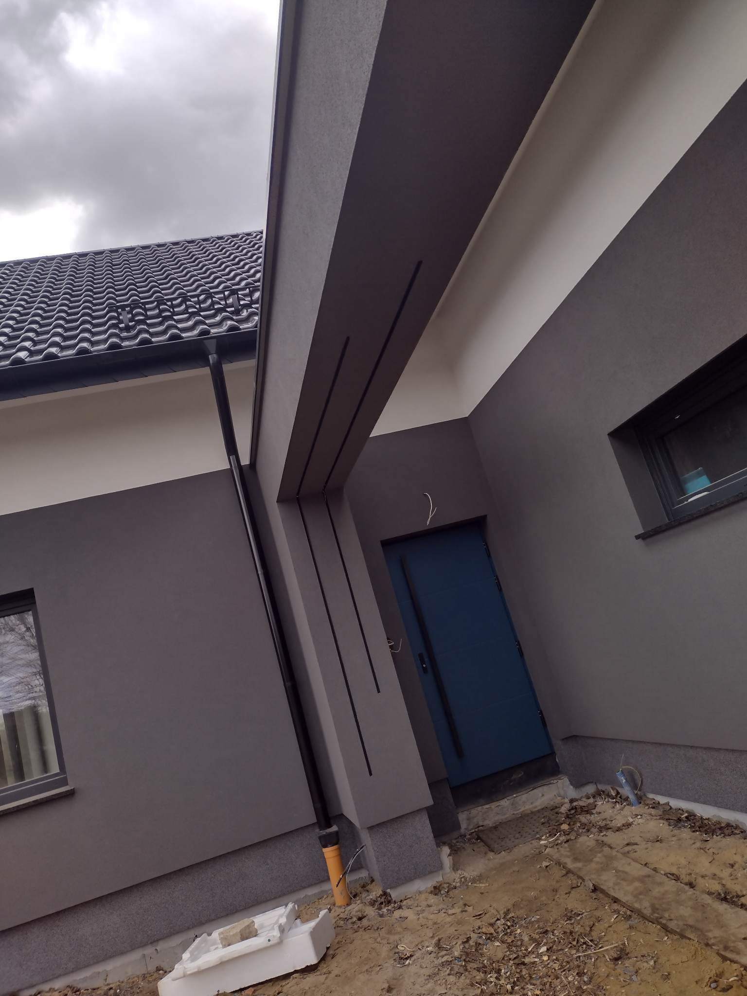 Szara elewacja domu z ciemną stolarką okienną i niebieskimi drzwiami wejściowymi, widoczny fragment dachu i rynna, teren wokół domu w trakcie prac wykończeniowych.