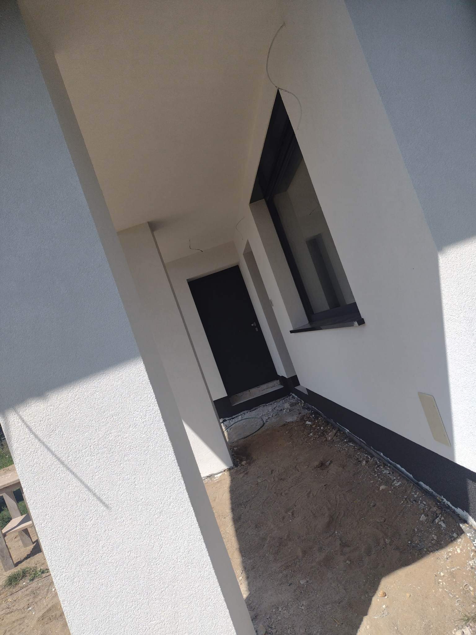 Nowo otynkowana elewacja domu z widocznym oknem i drzwiami wejściowymi, teren wokół w trakcie przygotowania.