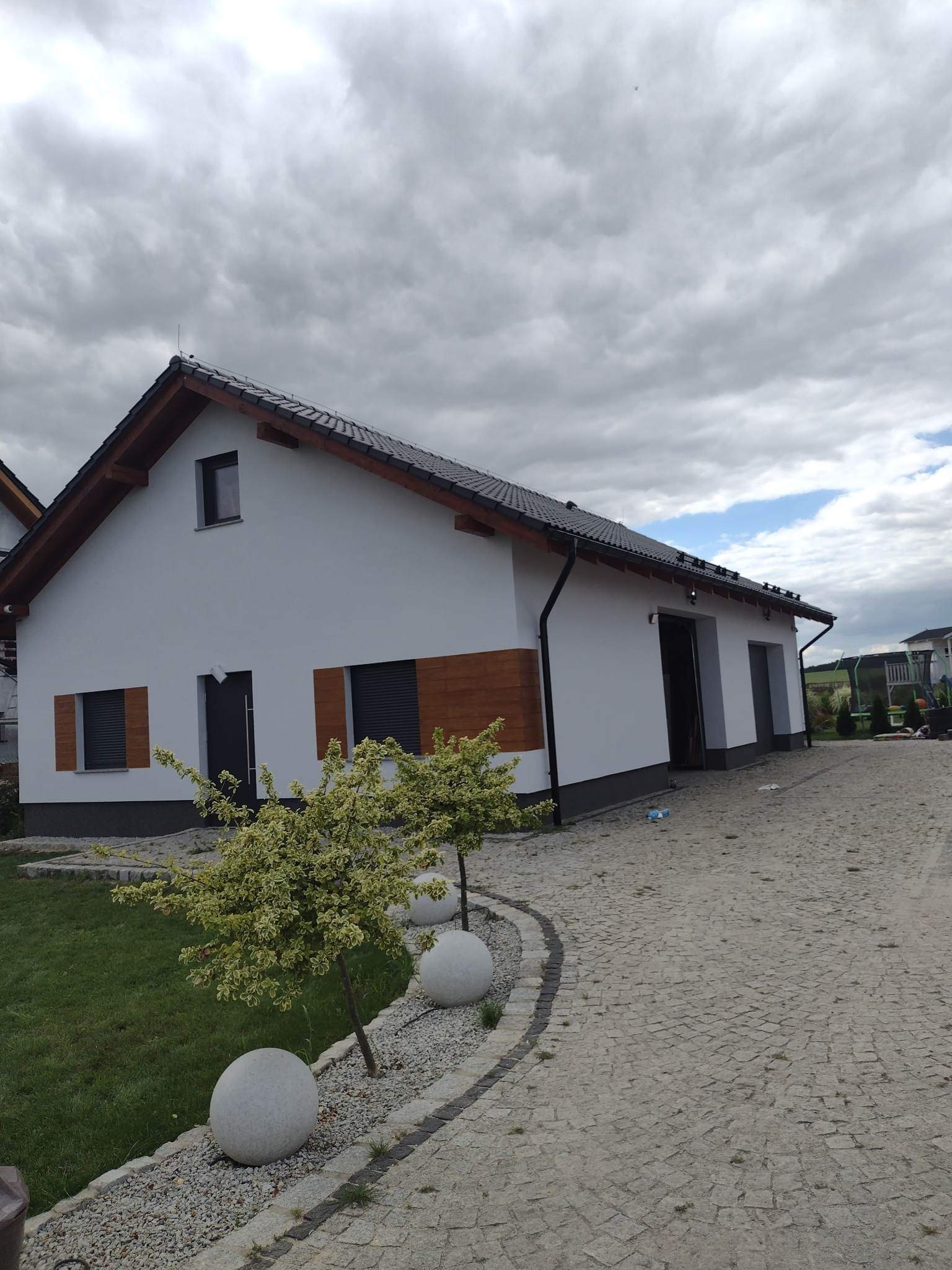 Biała elewacja domu jednorodzinnego z drewnianymi okiennicami i ciemną stolarką okienną oraz kostką brukową na podjeździe, widok w pochmurny dzień.