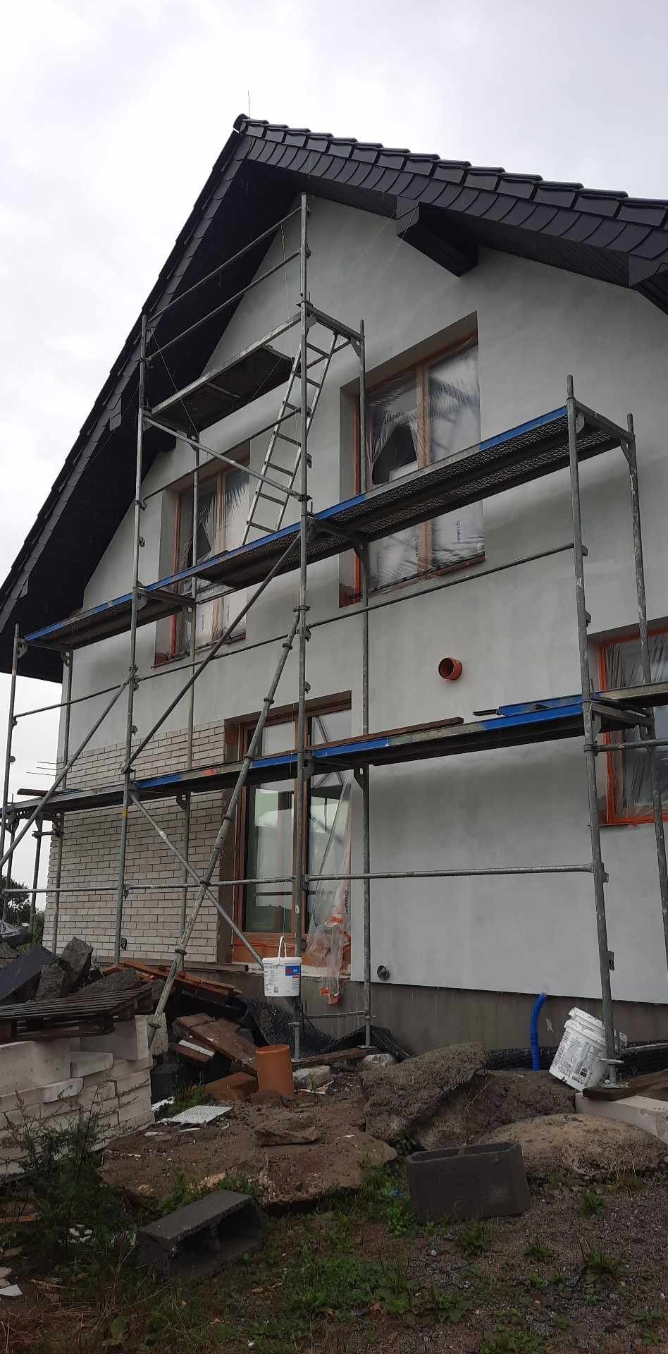 Dom jednorodzinny w trakcie prac elewacyjnych: ściany pokryte szarą warstwą, okna zabezpieczone folią, metalowe rusztowanie, widoczne materiały budowlane i wiadro z farbą u podstawy ściany.