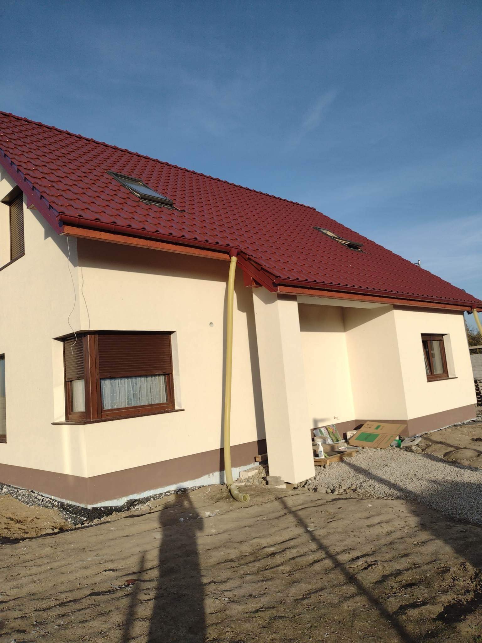Jasna elewacja domu jednorodzinnego z brązowymi oknami i czerwoną dachówką, widoczna rynna spustowa i teren wokół budynku w trakcie zagospodarowywania.