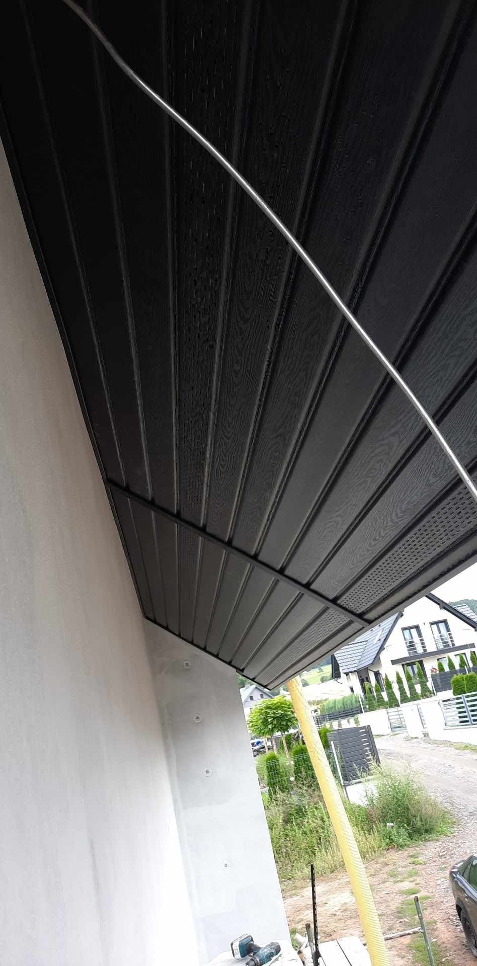 Zamontowana podbitka dachowa z ciemnego drewna na zewnątrz budynku, widoczna z dołu pod kątem. Na ścianie widoczne ślady po mocowaniach. W tle dom jednorodzinny i zieleń.