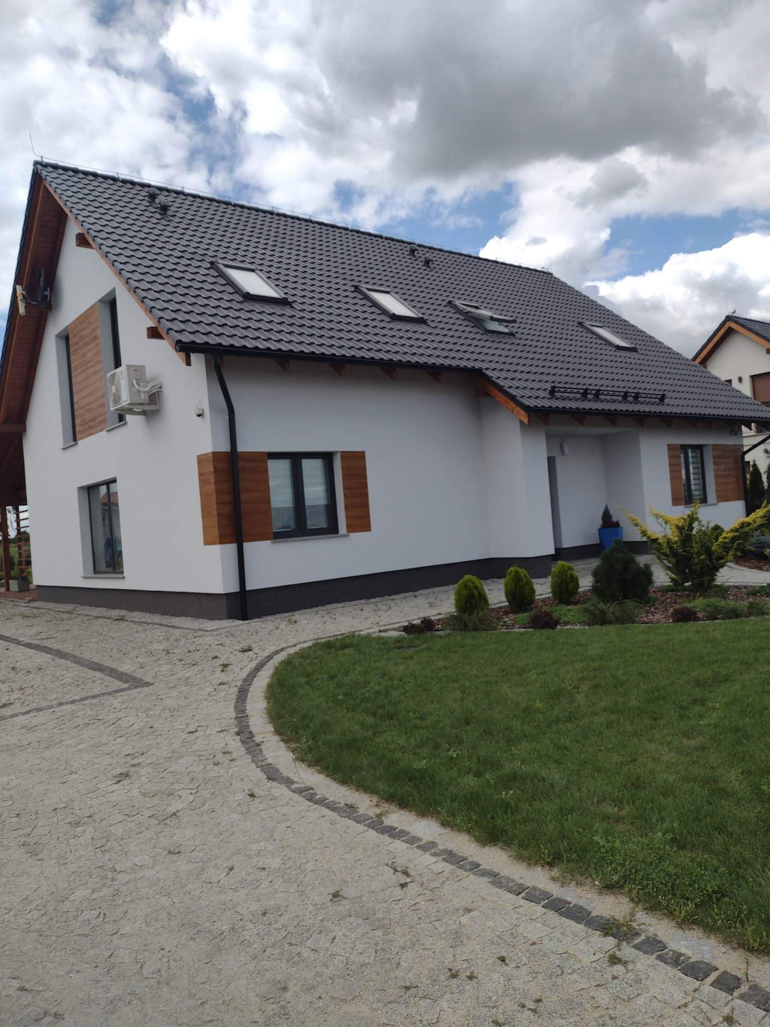 Biały dom jednorodzinny z ciemnoszarą dachówką, drewnianymi akcentami na elewacji i zadbanym trawnikiem oraz ścieżką z kostki brukowej.