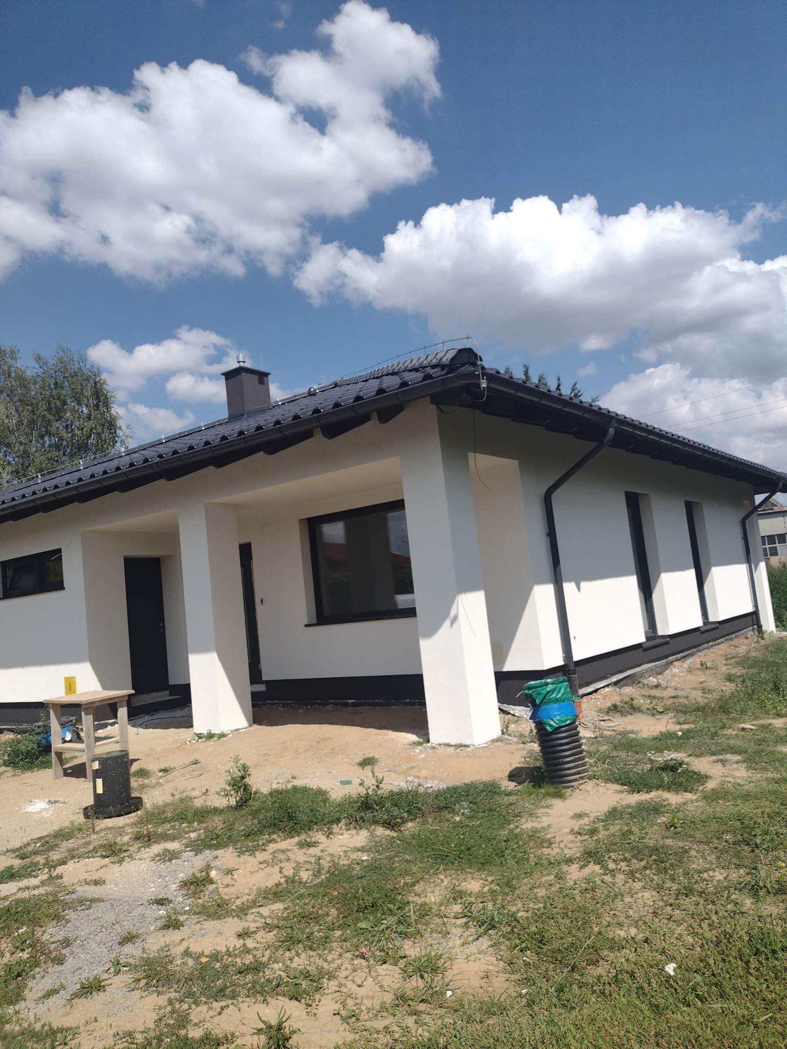 Świeżo pomalowany, parterowy dom z ciemną dachówką i stolarką okienną, widok z zewnątrz w słoneczny dzień, z widocznym fragmentem terenu przed domem i drewnianym stołkiem malarskim.