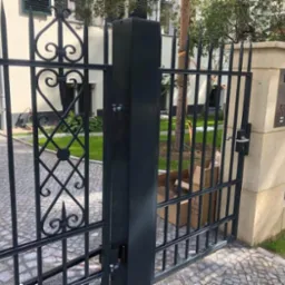 Otwarta, czarna brama kuta z dekoracyjnymi elementami w kształcie serc i grotów, zamontowana przy kamiennym słupku z numerem domu 53, widok na brukowany podjazd i trawnik.