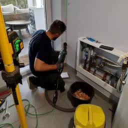 Hydraulik wierci otw&oacute;r w ścianie w pobliżu skrzynki z zaworami, używając wiertarki udarowej i lasera krzyżowego, obok stoi ż&oacute;łty odkurzacz przemysłowy i wiadro z resztkami gruzu.