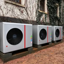 Trzy szare jednostki zewnętrzne z czerwonymi paskami, ustawione na betonowych podstawach przed budynkiem porośniętym bluszczem.