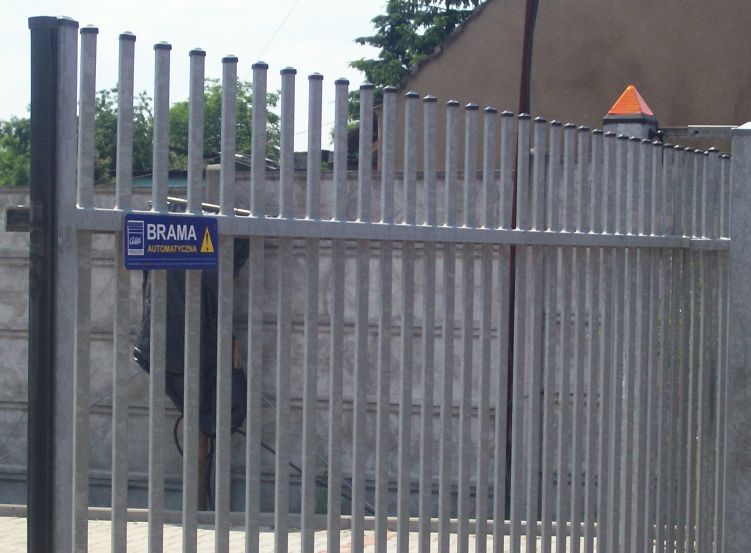 Szara metalowa brama automatyczna z pionowymi szczeblami i pomarańczowym ostrzegawczym stożkiem na szczycie, na tle betonowego muru i zieleni.