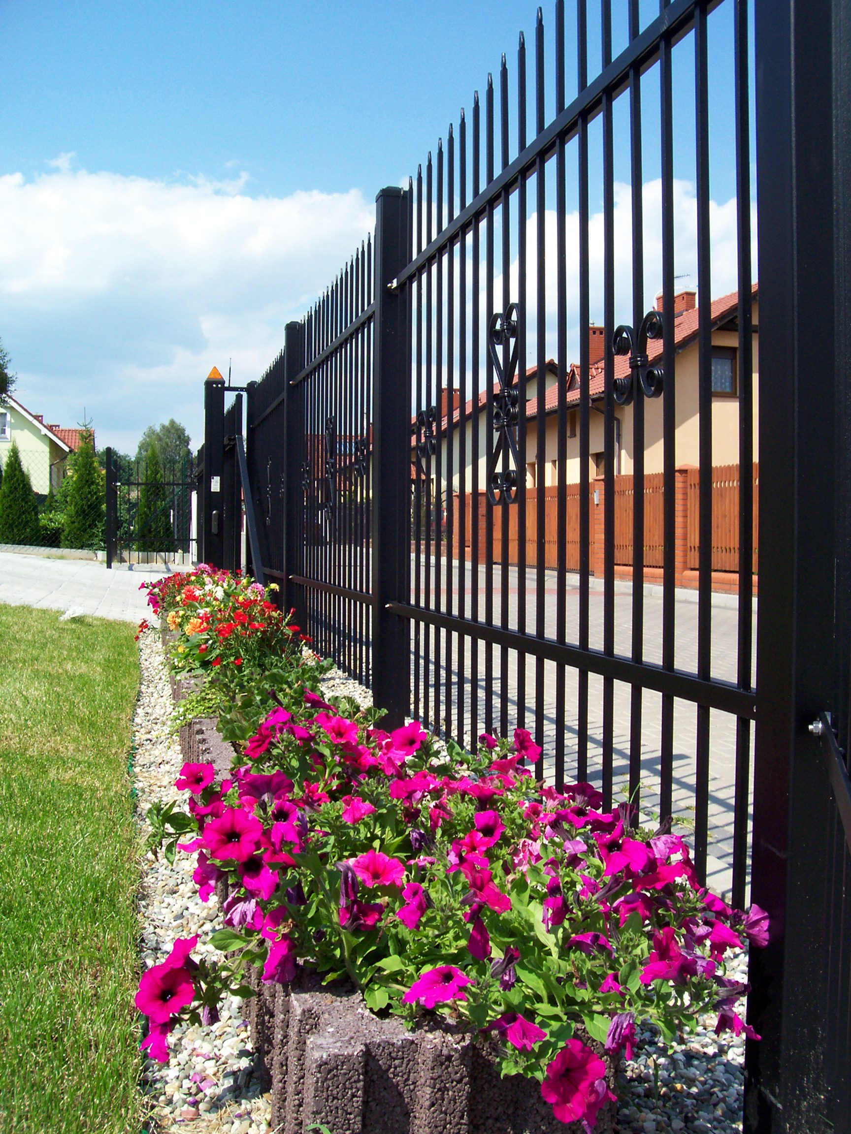 Czarne, metalowe ogrodzenie posesji z ozdobnymi elementami, widok z boku na rabatę kwiatową w betonowych obrzeżach, w tle domy jednorodzinne pod błękitnym niebem.