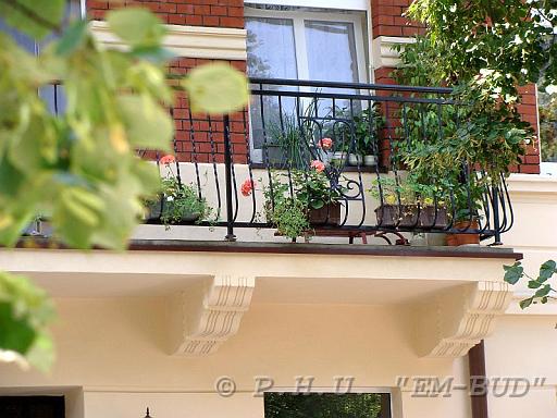 Kuty balkon z kwiatami w donicach, widok z dołu przez liście drzew, odnowiona elewacja budynku z cegły.