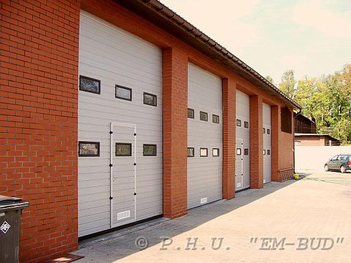 Budynek z cegły z czterema szarymi, segmentowymi bramami garażowymi z małymi okienkami, widoczne z zewnątrz, z lekkim efektem rybiego oka.