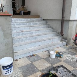 Baz-Bud - Wyłożone szarym gresem schody zewnętrzne w trakcie remontu, widoczny kubeł z zaprawą PROGOLD, szlifierka kątowa, dłuto i fragmenty płytek na szachownicowej posadzce.