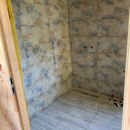 Baz-Bud - Niedokończona łazienka z ułożonymi płytkami ściennymi w kwiatowy wzór i podłogowymi imitującymi drewno, widoczne elementy instalacji hydraulicznej i narzędzia glazurnicze.