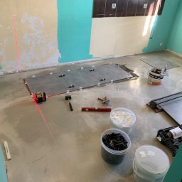Baz-Bud - Układanie szarych płytek podłogowych w pomieszczeniu z pomalowanymi na biało-turkusowo ścianami, widoczny laser krzyżowy wyznaczający poziom, poziomica, młotek gumowy, wiaderka z krzyżykami...