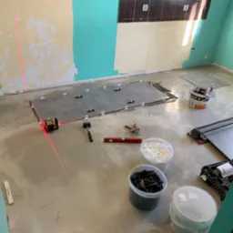 Układanie szarych płytek podłogowych w pomieszczeniu z pomalowanymi na biało-turkusowo ścianami, widoczny laser krzyżowy wyznaczający poziom, poziomica, młotek gumowy, wiaderka z krzyżykami...