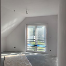 Baz-Bud - Pokój na poddaszu w trakcie prac wykończeniowych; okno zabezpieczone folią, ściany świeżo pomalowane na biało, widoczne gniazdka elektryczne bez osprzętu, na podłodze płyta OSB.