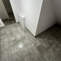 Baz-Bud - Świeżo położone, szare płytki podłogowe w pomieszczeniu ze skośnym sufitem, widoczny fragment buta roboczego z zabrudzeniami, biała szafka w rogu, ślady po pracach budowlanych w tle.