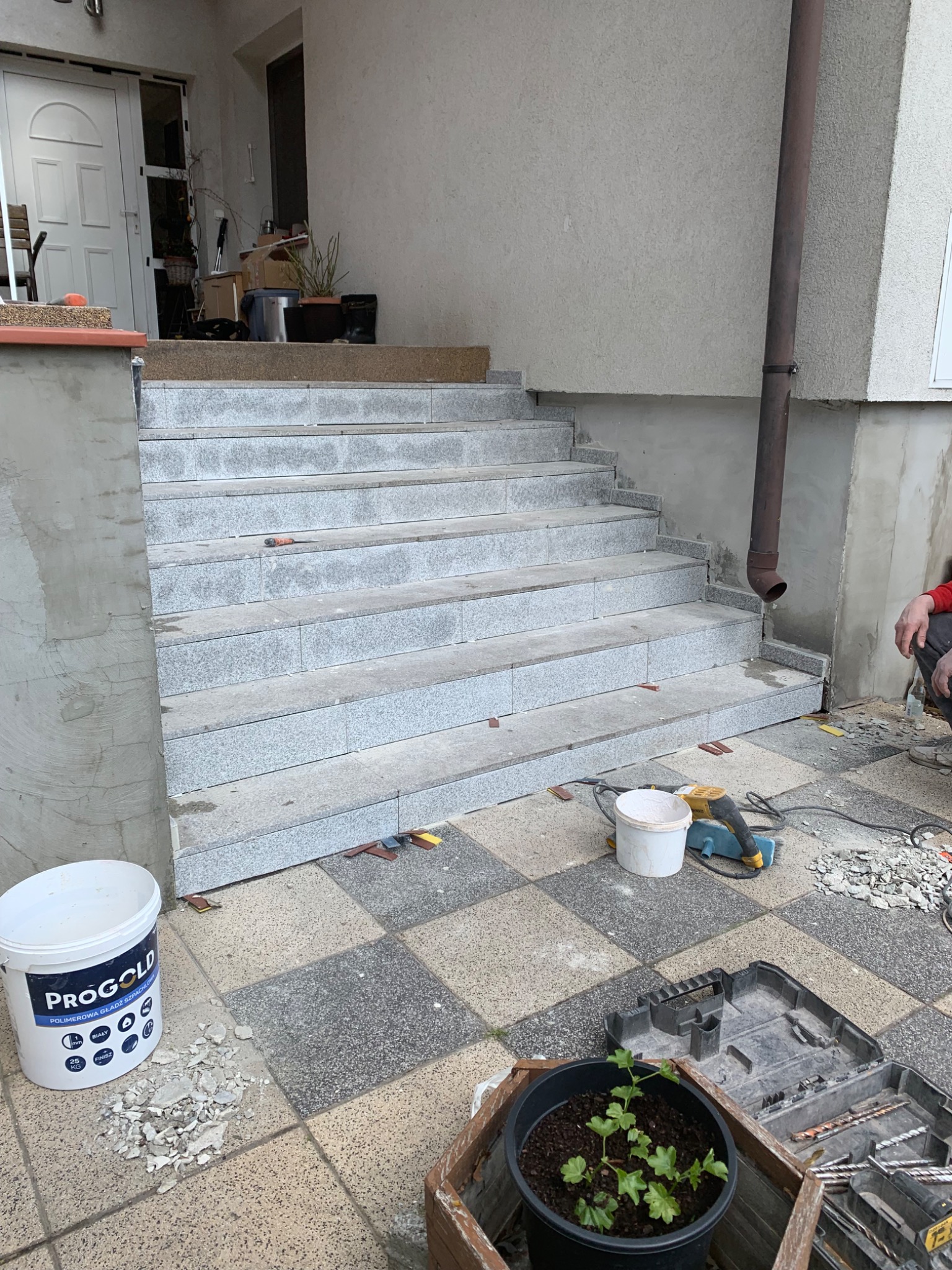 Wyłożone szarym gresem schody zewnętrzne w trakcie remontu, widoczny kubeł z zaprawą PROGOLD, szlifierka kątowa, dłuto i fragmenty płytek na szachownicowej posadzce.